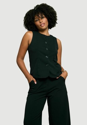 Donna con capelli ricci che indossa un gilet nero senza maniche con bottoni e pantaloni neri, in piedi con le mani nelle tasche davanti a uno sfondo semplice.