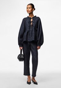 Blusa blu marino a texture con maniche a sbuffo, con laccio anteriore, abbinata a pantaloni coordinati e una borsa nera piccola. Calzature includono tacchi neri.