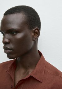 Rust-colored linen shirt with a collar, featuring a textured fabric and a relaxed fit, worn by a person in profile against a neutral background.