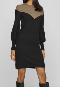 Robe tricot noir avec un design de poitrine marron, manches bouffantes longues et poignets côtelés. Longueur genou avec une coupe droite, fabriquée à partir d'un matériau doux.