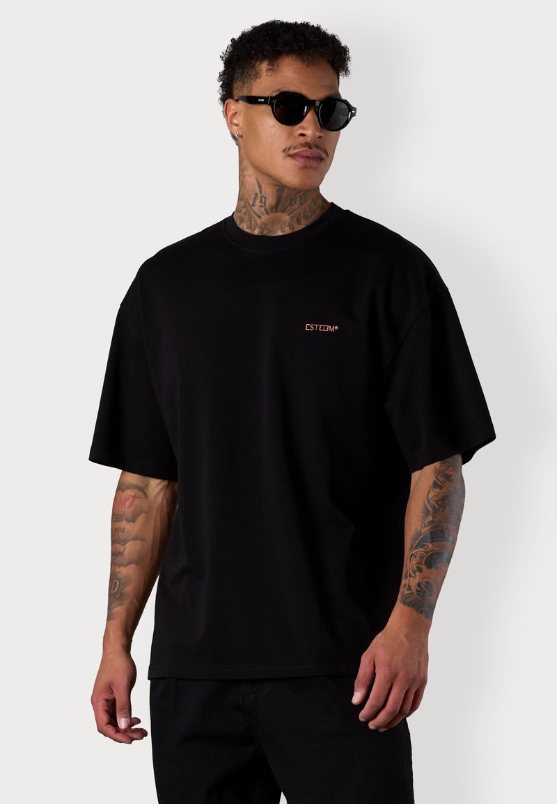 Schwarzes oversized T-Shirt aus Baumwolle mit kurzen Ärmeln und einem kleinen orangefarbenen ESTIM-Logo auf der linken Brust. Getragen mit einer schwarzen Sonnenbrille.