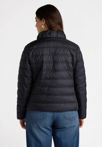 Chaqueta acolchada negra con un patrón de rombos, cuello alto y un diseño corto. Lleva puesta sobre jeans de denim, con una textura lisa y brillante.