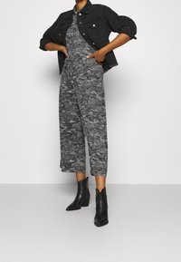 Svart kort jacka lagd över en mönstrad jumpsuit i grått och vitt, med vida ben och svarta klackstövlar i ankellängd.