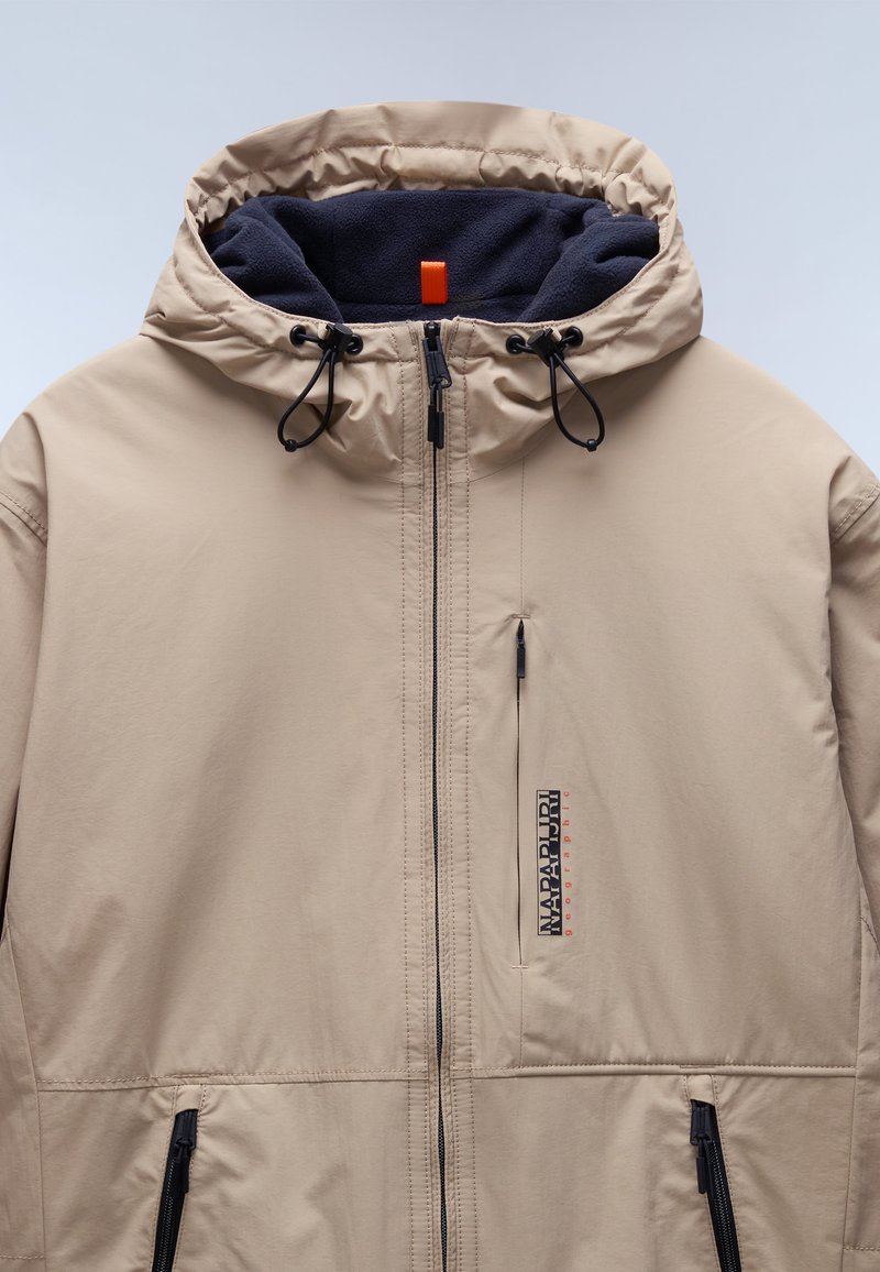 Beige gewatteerde jas met een gladde stof, voorzien van een marineblauwe fleece voering, een ritssluiting aan de voorkant, trekkoorden en een logo patch op de borst.