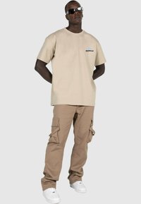 Beige bomull t-skjorte med et grafisk logo på brystet, kombinert med lysebrune cargobukser med flere lommer og hvite sneakers.