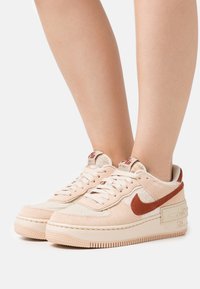 Light beige sneakers with a textured upper and rust-colored swoosh. Thick rubber sole and lace-up design with padded ankle collar.