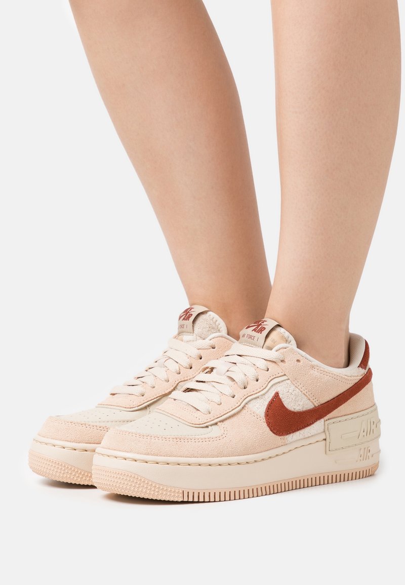 Light beige sneakers with a textured upper and rust-colored swoosh. Thick rubber sole and lace-up design with padded ankle collar.