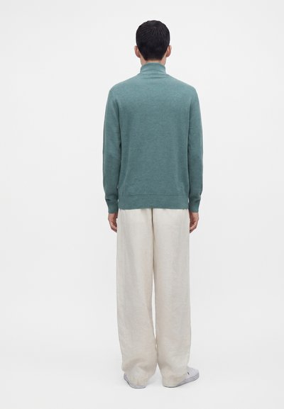 Personne debout, dos face à l’avant, portant un pull à col roulé bleu sarcelle, un pantalon blanc ample et des chaussures blanches sur fond blanc uni.