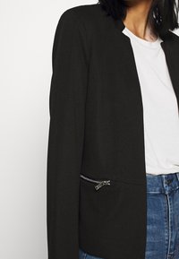 Černý blejzr s texturovaným materiálem. Má zkrácený střih a stříbrnou kapsu na zip v přední části. Nosený přes bílou blůzu.