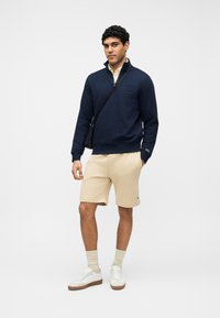 Homme portant un sweat-shirt bleu marine à demi-fermeture éclair, un short beige, des chaussettes hautes, des baskets blanches et un sac bandoulière noir, debout devant un fond blanc.