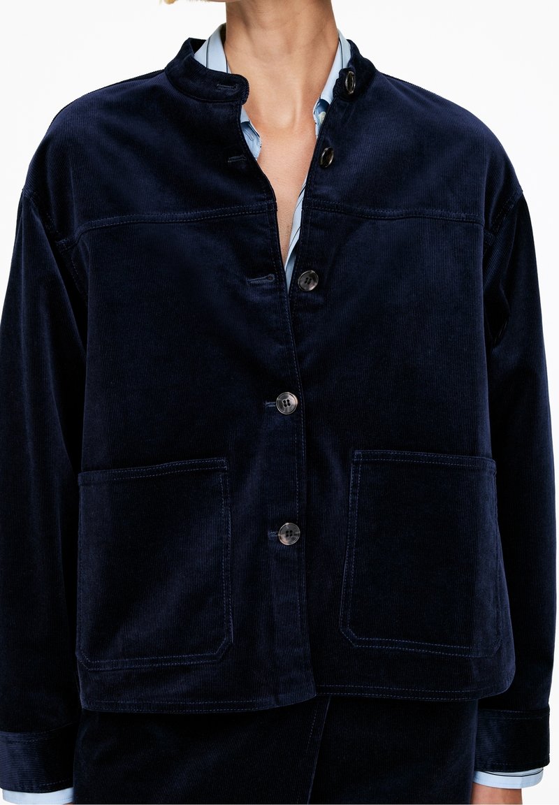 Veste en velours côtelé bleu foncé avec fermeture à boutons, grandes poches avant et col montant portée sur une chemise bleu clair.