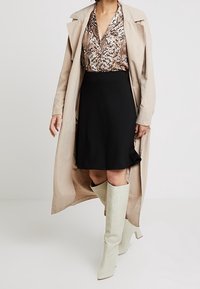 Un trench-coat beige sur un chemisier marron à motifs, une jupe noire évasée et des bottes vert menthe montantes. Présente un décolleté en V et un tissu fluide.