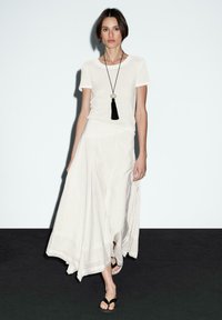 Gonna maxi bianca in lino con orlo ampio e fluido, abbinata a una t-shirt bianca aderente e una collana con nappine nera. Sandali neri completano il look.