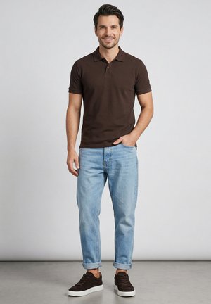 Usmívající se muž stojí, oblečený v tmavě hnědém polotriku, světle modrých ohrnutých džínách a tmavě hnědých teniskách na jednoduchém pozadí.