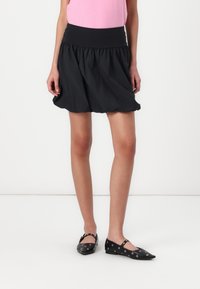 Jupe noire évasée avec une taille élastique froncée, fabriquée en matériau léger. Associée à des ballerines noires avec des découpes circulaires.