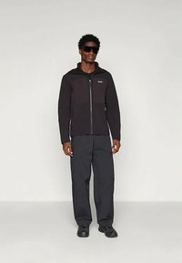 Black zip-up jacket with a hood and contrasting panel, worn with dark trousers and black shoes, all set against a neutral background.