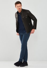 Zwarte leren jas met geborduurde schouderdetails, marineblauw shirt, aansluitende teal broek en zwarte schoenen. Staande pose, neutrale achtergrond.