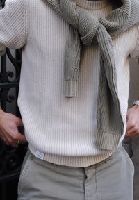Pull beige clair en tricot avec texture côtelée et une grande écharpe tricotée vert olive drapée autour du cou. Pantalon gris avec une étiquette visible.