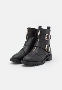 Bottes à cheville noires en cuir lisse, avec des accessoires couleur or, deux sangles décoratives et des fermetures éclair latérales. Semelle en caoutchouc épaisse.