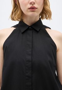 Γυναίκα με ανοιχτόχρωμο δέρμα και κυματιστά ξανθά μαλλιά που φοράει αμάνικο μαύρο μπλουζάκι με γιακά και λεπτομέρειες κοψίματος στους ώμους.