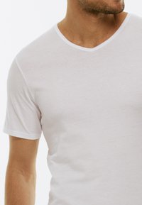 T-shirt bianco a collo a v con maniche corte, realizzato in tessuto morbido. Presenta una sottile texture e un bordo bianco attorno al collo.