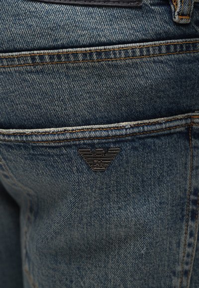 Φθαρμένα μπλε τζην ξεχωρίζουν με μια μικρή, μαύρη ανάγλυφη λεπτομέρεια λογότυπου στην πίσω τσέπη και αντίθετη καφέ ραφή.