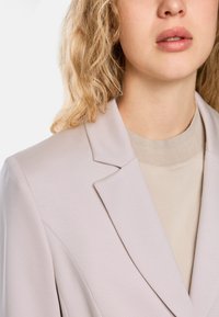 Hellgrauer Blazer mit strukturiertem Finish, ausgestattet mit einem Ein-Knopf-Verschluss und Reverskragen, über einem beigen Pullover.