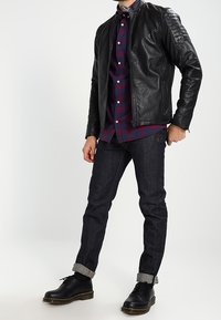 Black leather jacket with ribbed shoulder details, red and blue checkered shirt, dark denim jeans, cuffed hems, and black lace-up boots.