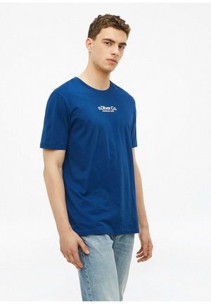Młody mężczyzna w niebieskim T-shircie "s.Oliver Co." i jasnoniebieskich dżinsach, stojący na jednolitym białym tle.