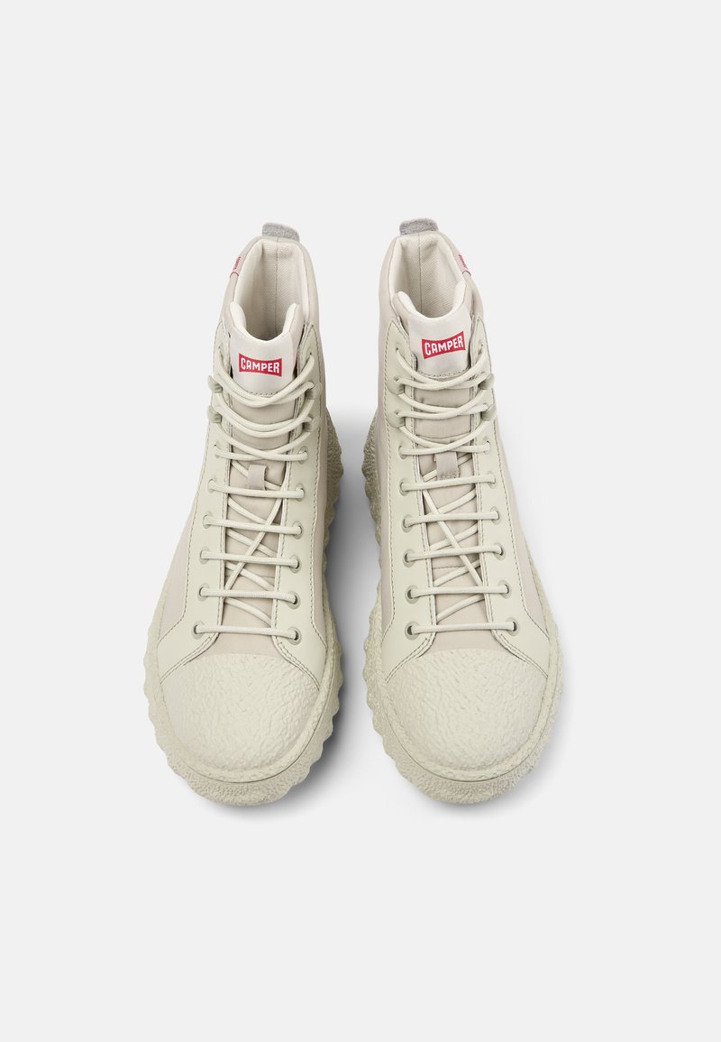 Zapatillas altas en beige claro, con suelas de goma texturizadas, un upper liso con cierres de cordones y un parche del logo rojo en la lengüeta.