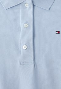 Lyseblå polo skjorte laget av teksturert stoff med krage og tre hvite knapper. Har et lite rødt, hvitt og svart logo.