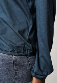 Dunkelblaugrüne, leichte Bluse mit langen Ärmeln, gerafftem Saum und Knopfdetails. Weiche Textur und glattes Material. Kombiniert mit grauen Jeans.