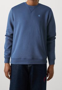Felpa blu con collo rotondo e polsini a costine. Caratterizzata da un piccolo logo sul petto e un tessuto morbido e texture. Indossata con jeans scuri.