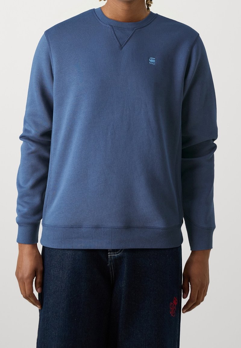 Felpa blu con collo rotondo e polsini a costine. Caratterizzata da un piccolo logo sul petto e un tessuto morbido e texture. Indossata con jeans scuri.