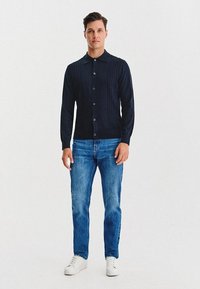 Mężczyzna stoi w czarnym swetrze zapinanym na guziki, niebieskich dżinsach i białych sneakersach na tle jednolitego białego tła.