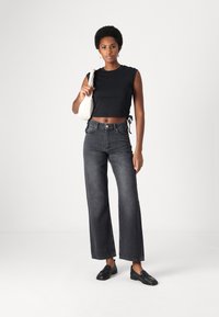 Black cropped top with tied sides, paired with high-waisted, wide-leg dark gray jeans. Accessories include a light-colored handbag and black loafers.