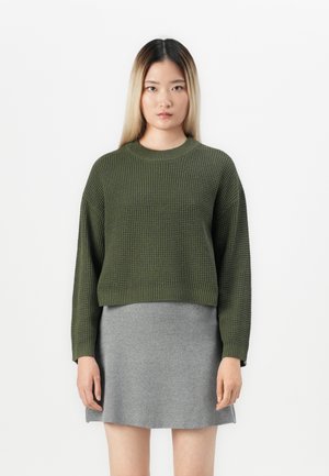 Kobieta stoi na białym tle, ubrana w oliwkowozielony sweter z dzianiny i szarą spódnicę, patrząc prosto przed siebie z neutralnym wyrazem twarzy.