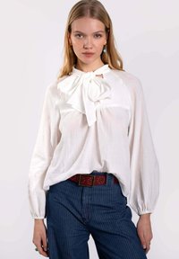 Blouse blanche à manches longues avec un col à nœud, fabriquée en tissu léger. Associée à un jean rayé bleu foncé et une ceinture rouge à pois.