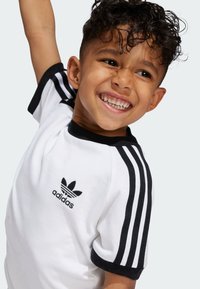 Biela tričko s krátkymi rukávmi, čiernym golierom a logom Adidas na hrudi, s čiernymi pruhmi na rukávoch. Hladká bavlnená textúra.