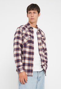 Camisa de franela con un patrón de cuadros en rojo, azul y blanco, dos bolsillos en el pecho con detalles de botón y un corte relajado. Material de algodón.