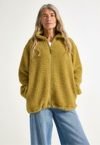 Donna con capelli lunghi indossa una giacca in pile color senape, accogliente, e jeans azzurri larghi, in piedi davanti a uno sfondo chiaro, con le braccia rilassate.