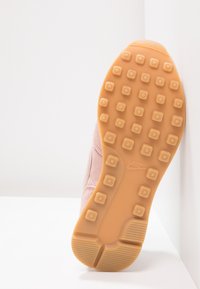 Růžová semišová podrážka tenisky s hnědou gumovou podešví, která má čtvercové vzory pro lepší přilnavost a logo Nike. Třídílný design podrážky s zaoblenými hranami.