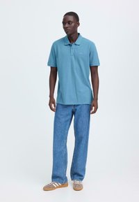 Polo turquoise avec col, manches courtes et détail logo, associé à un jean denim bleu et des baskets grises avec des rayures blanches.
