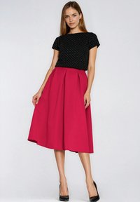 Kobieta w czarnej bluzce z krótkim rękawem w białe kropki oraz jasno różowej, midi spódnicy, stojąca na tle jasnego, jednolitego tła.