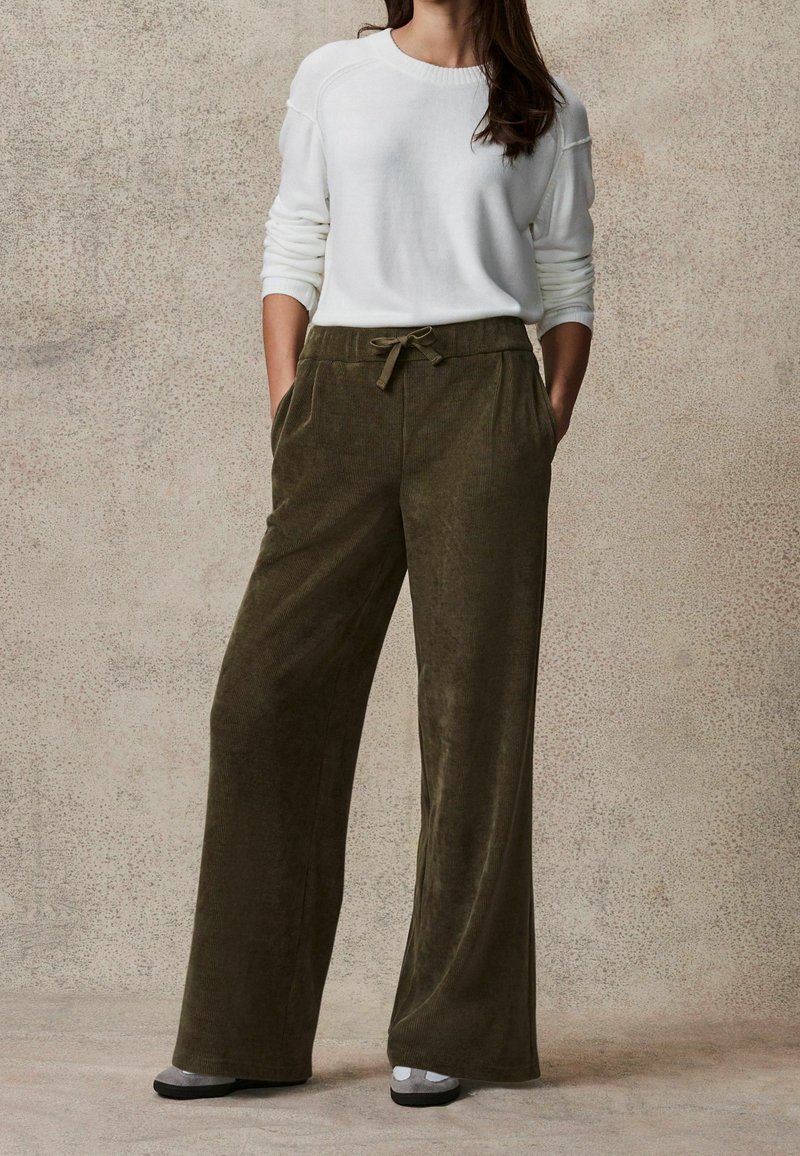 Oliwkowe kordowe spodnie o szerokich nogawkach z przednimi kieszeniami i regulowanym paskiem w talii, połączone z białym swetrem z długim rękawem.