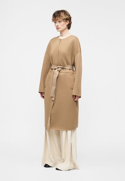 Γυναίκα με κοντά μαλλιά που φοράει μπεζ παλτό με ζώνη πάνω από φαρδιά κρεμ παντελόνια και μαύρα παπούτσια, στέκεται μπροστά σε απλό λευκό φόντο.