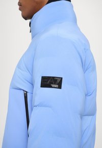 Light blue puffer jacket with black zipper and EA7 logo patch on the shoulder; features a high collar and smooth texture.