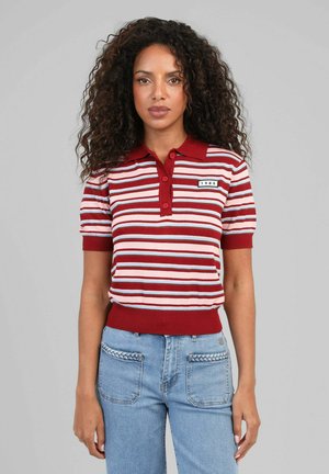 Femme aux cheveux bouclés portant un polo rayé rouge, blanc et rose et un jean bleu clair avec des détails tressés sur les poches.