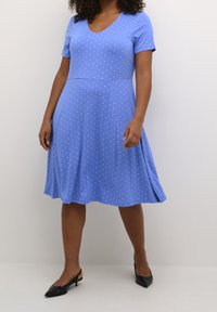 Kortærmet, V-hals kjole i lyseblåt stof med hvide polkaprikker, A-linjeform og knælængde. Sorte hæle supplerer outfittet.