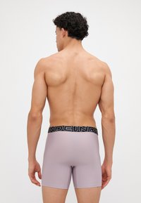 Helle lila Boxer-Shorts mit einem schwarzen elastischen Bund, der das Logo "UNDER" trägt. Glattes Material, figurbetontes Design, Ansicht von hinten.